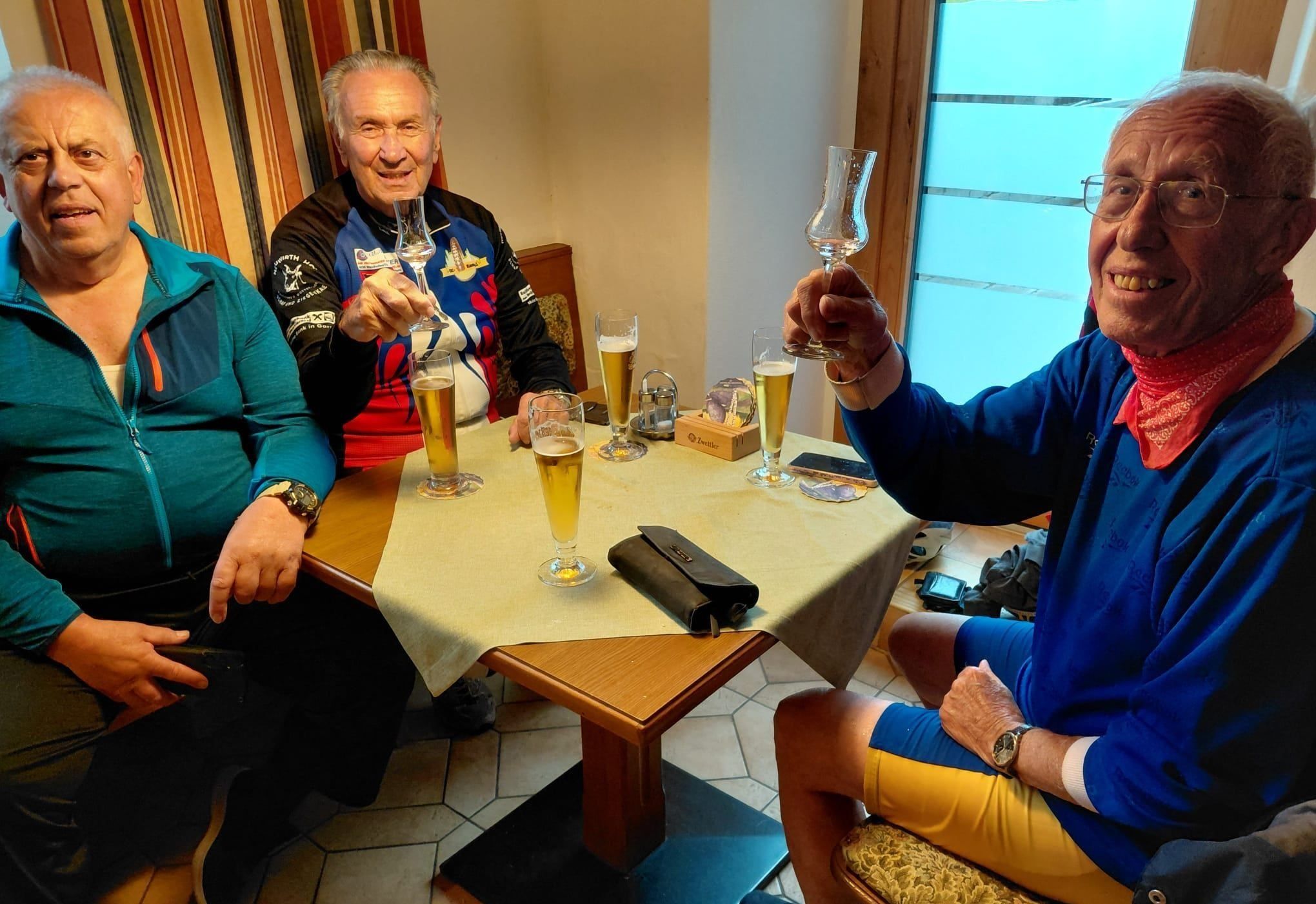 Drei ältere Männer stoßen mit Biergläsern in einem Raum an. Ein Mann trägt eine Uhr und ein Armband. Ein Tisch hat Biergläser, eine Geldbörse und eine Holzbox.