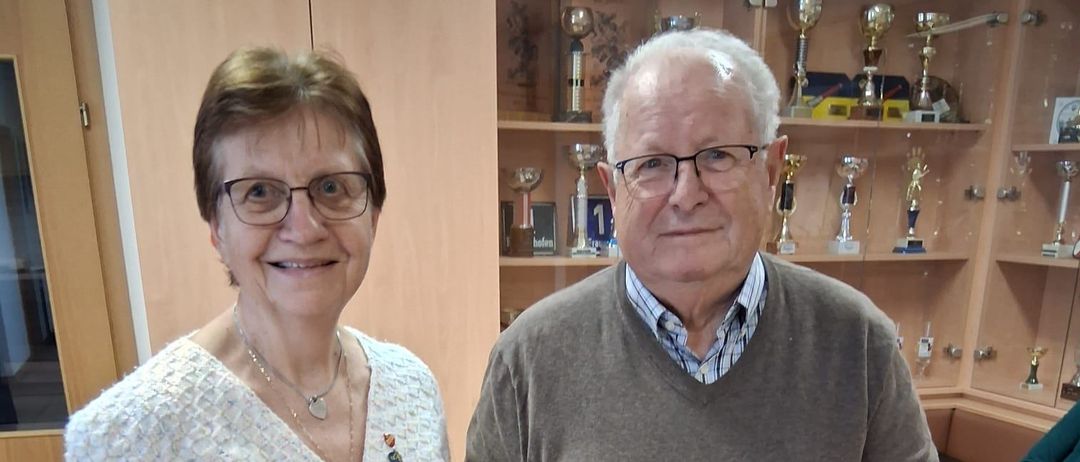 Ein älteres Paar steht in einer Innenraumszene und lächelt. Der Mann hält eine Tasche, und hinter ihnen befinden sich Regale mit Trophäen. Rechts steht ein Tisch mit Tassen und einer Serviette.