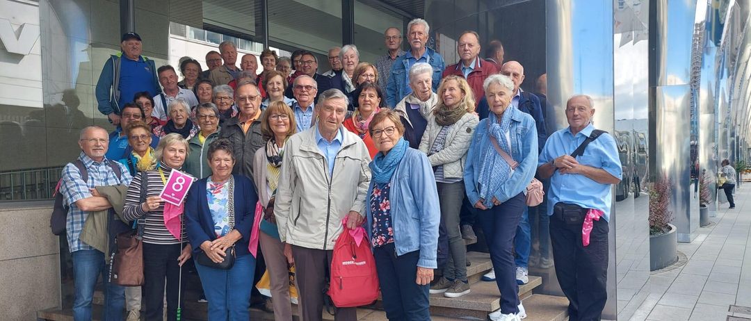 Eine Gruppe von Menschen unterschiedlichen Alters, in Freizeitkleidung gekleidet, steht auf den Stufen eines Gebäudes und hält teilweise Plakate. Sie lächeln und posieren für ein Foto.
