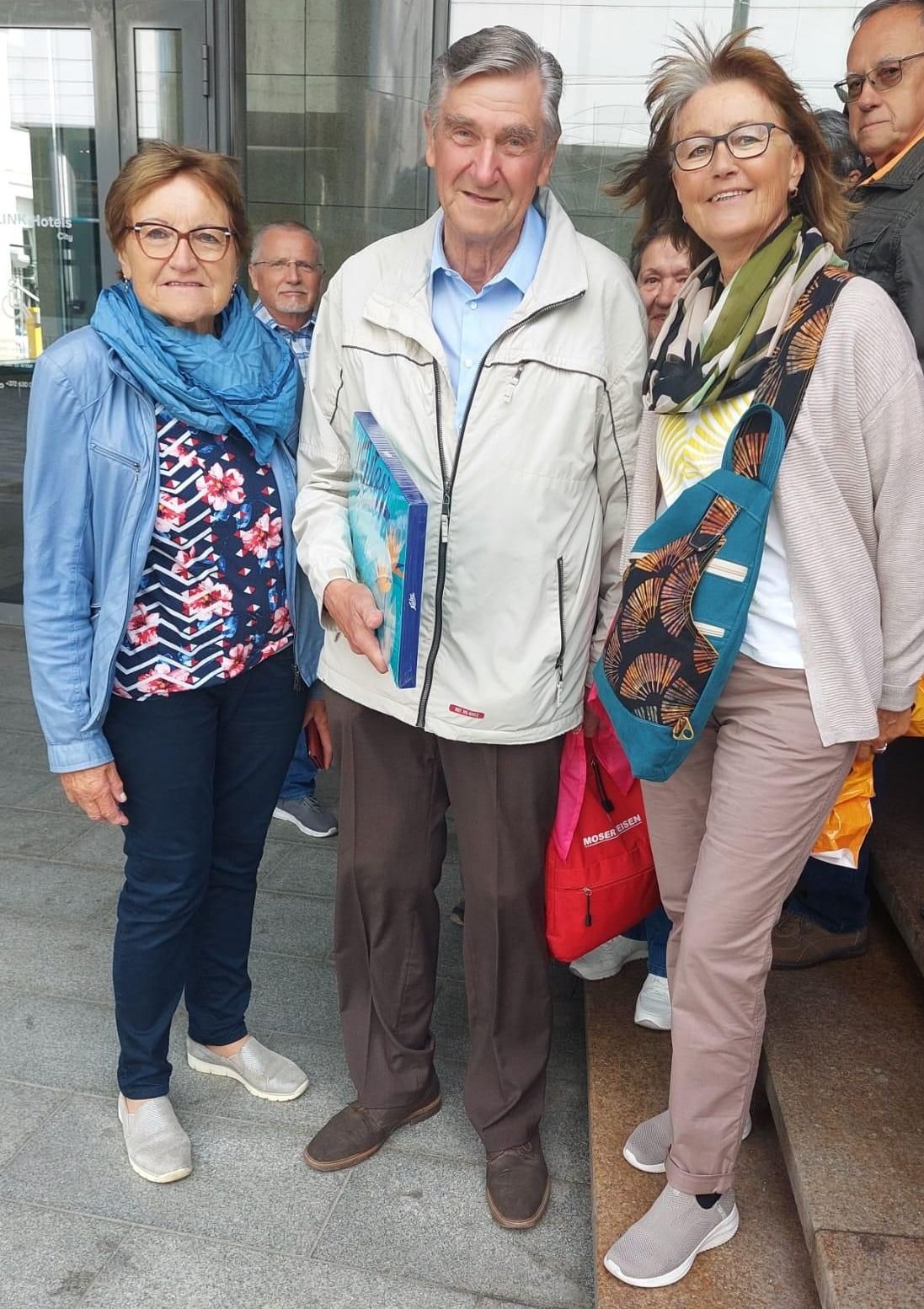 Drei Menschen stehen auf Stufen; ein Mann hält ein blaues Buch, eine Frau trägt einen blauen Schal und eine andere Frau trägt eine rote Tasche.