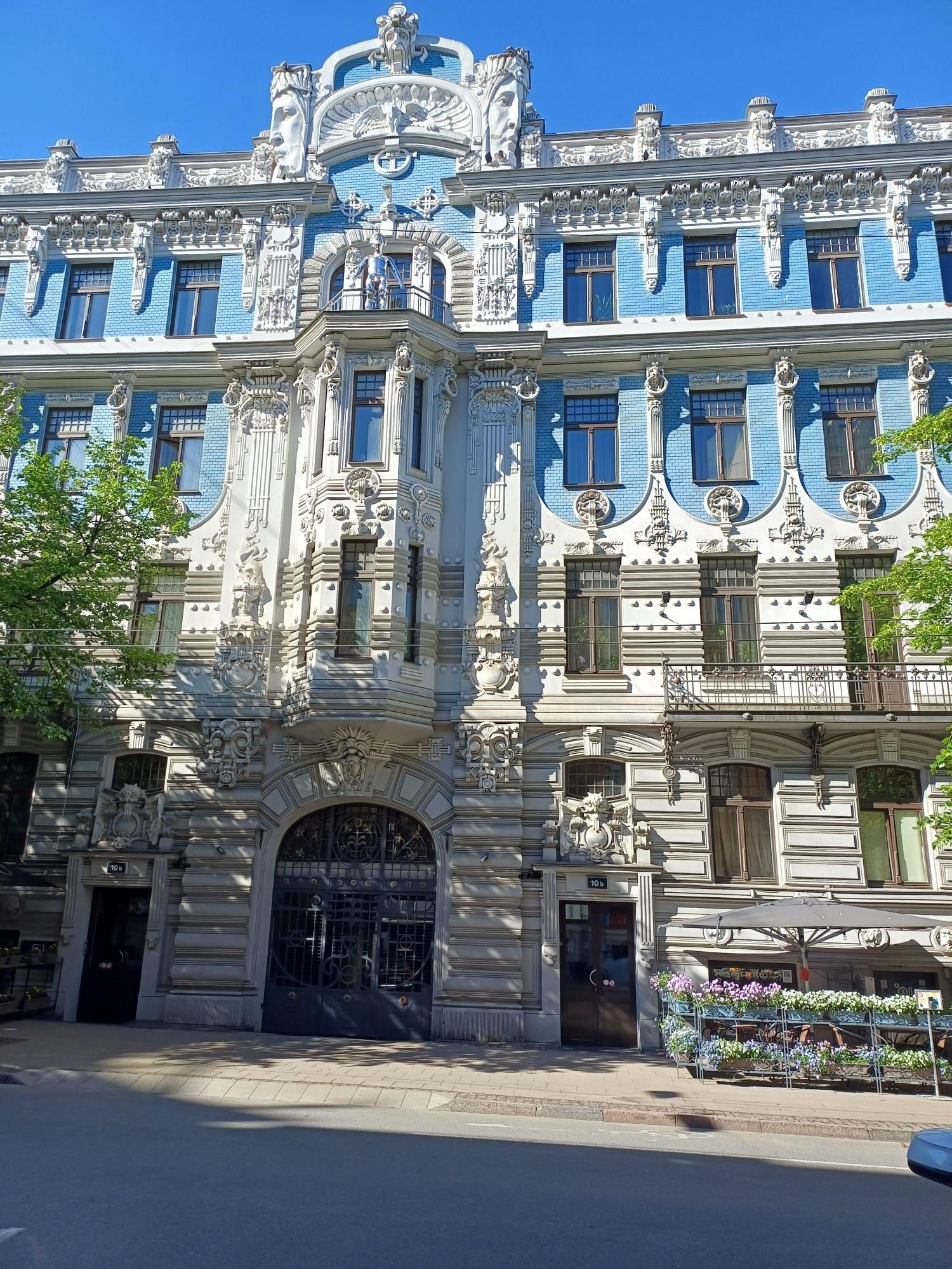 Ein großes, reich verziertes Gebäude mit blauen Kacheln und weißen Umrandungen, vielen Fenstern und dekorativen Skulpturen. Unten befindet sich ein offener Eingang mit einem Tor. Rechts gibt es einen kleinen Bereich mit Pflanzen und einem Schirm.