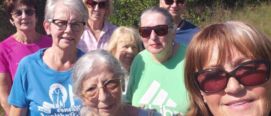 Eine Gruppe älterer Frauen in passenden T-Shirts lächelt und posiert für ein Foto im Park. Sie tragen Sonnenbrillen und stehen vor Bäumen.