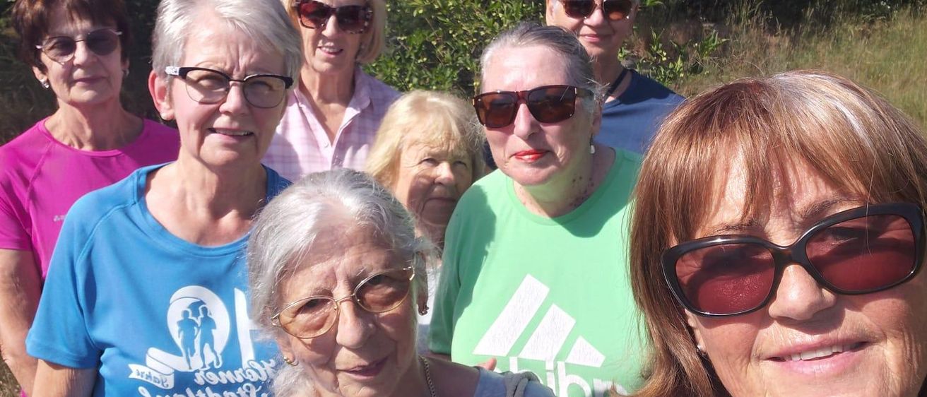 Eine Gruppe älterer Frauen in passenden T-Shirts lächelt und posiert für ein Foto im Park. Sie tragen Sonnenbrillen und stehen vor Bäumen.