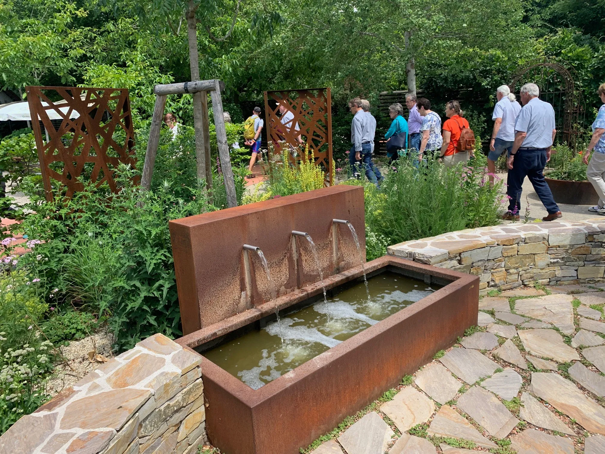 Eine Gruppe von Menschen geht in einem Garten mit einem Wasserbrunnen aus rostfreiem Metall herum. Der Brunnen hat drei Wasserspeier. Der Garten ist von Bäumen und Pflanzen umgeben.