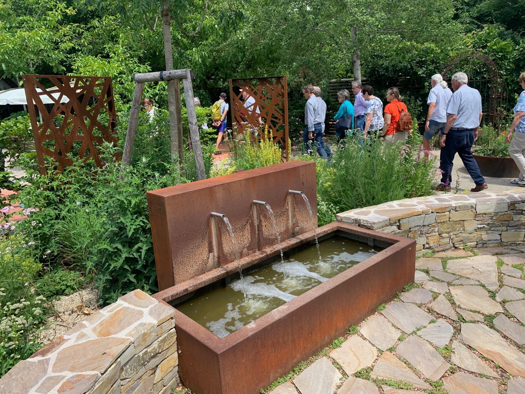 Eine Gruppe von Menschen geht in einem Garten mit einem Wasserbrunnen aus rostfreiem Metall herum. Der Brunnen hat drei Wasserspeier. Der Garten ist von Bäumen und Pflanzen umgeben.