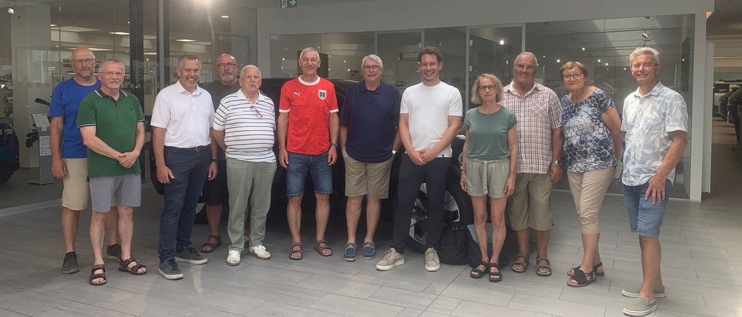 Eine Gruppe von Menschen, sowohl Männer als auch Frauen, steht in einem Ausstellungsraum. Einige lächeln, andere sehen ernst aus. Der Mann in Rot trägt ein Fußballtrikot.