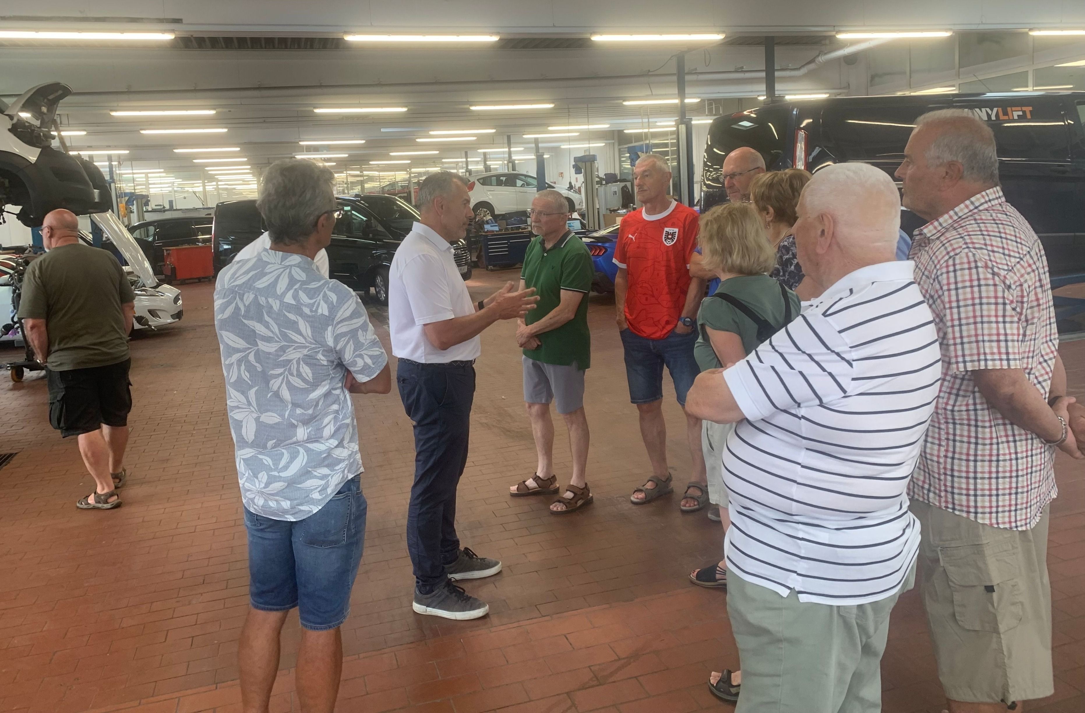 Eine Gruppe älterer Erwachsener unterhält sich in einem Autohaus. Einige tragen Brillen und Turnschuhe, während andere Sandalen tragen. Ein rotes Auto ist in der Nähe geparkt.