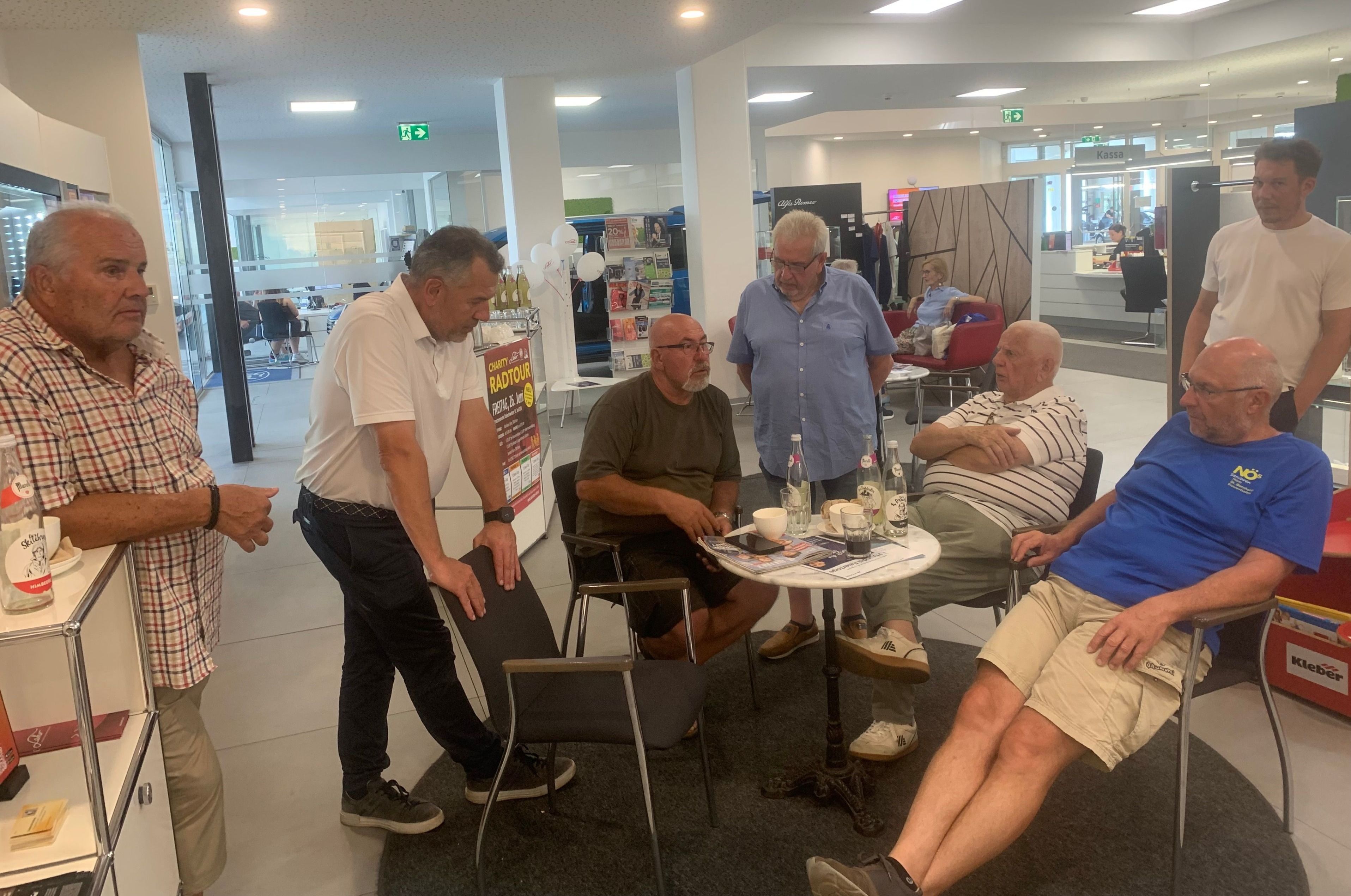 Fünf Männer sind in einer Lounge versammelt. Ein Mann steht bei einem Stuhl, ein anderer sitzt und drei weitere stehen. Sie trinken Getränke.
