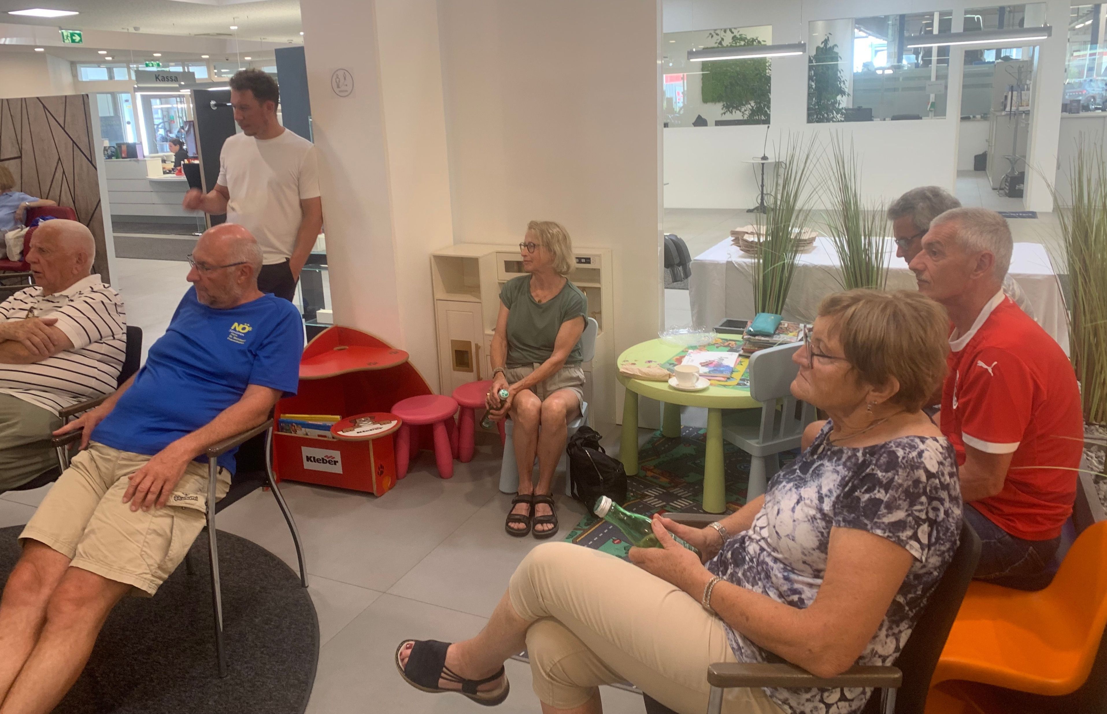 Eine Gruppe von Menschen sitzt um einen Tisch. Eine ältere Frau hält eine grüne Flasche. Ein Mann steht im Hintergrund. Stühle und Hocker sind um sie herum.