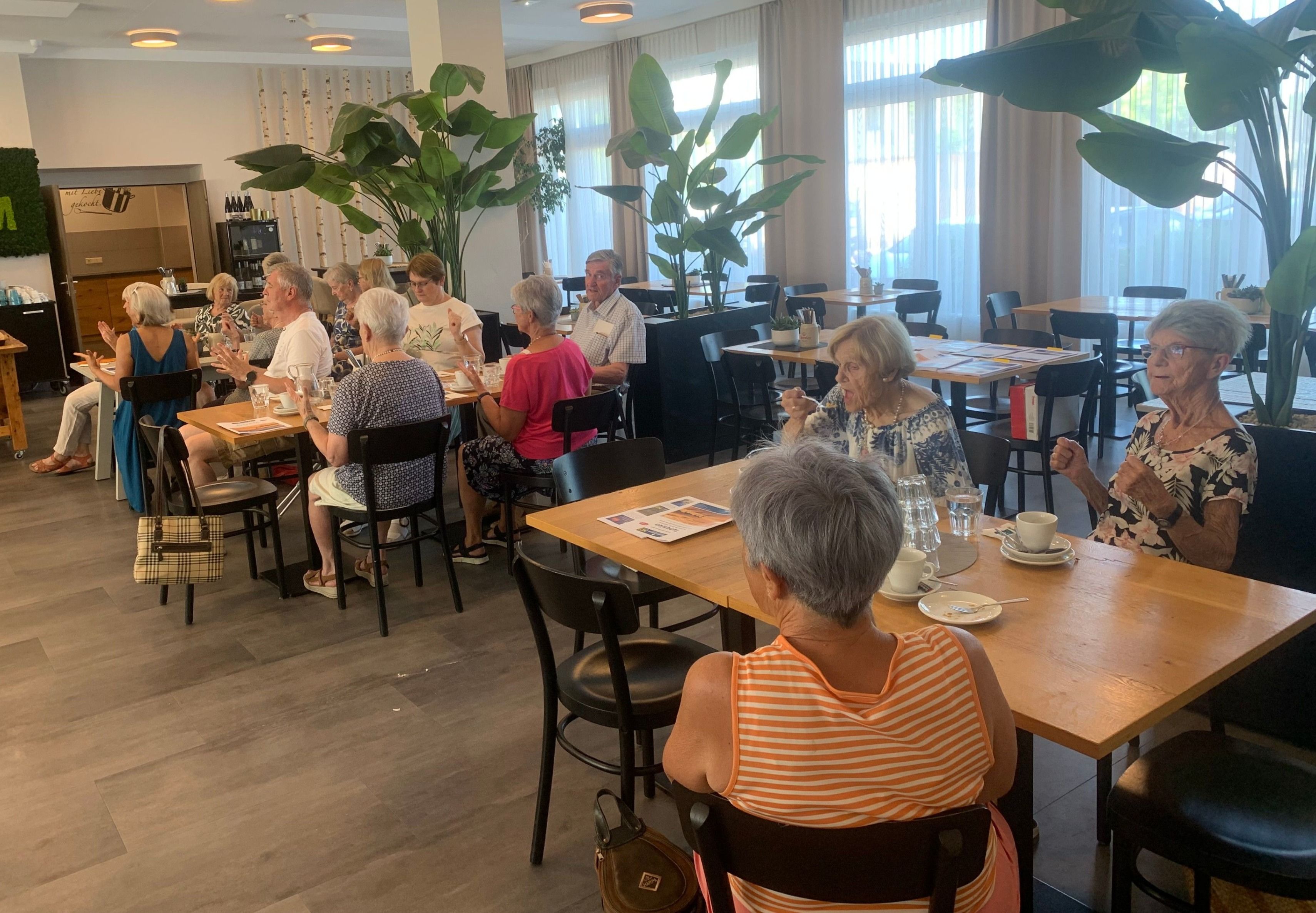 Mehrere ältere Menschen sitzen an Tischen in einem Esszimmer mit Pflanzen, Holzböden und großen Fenstern. Einige unterhalten sich, während andere essen.
