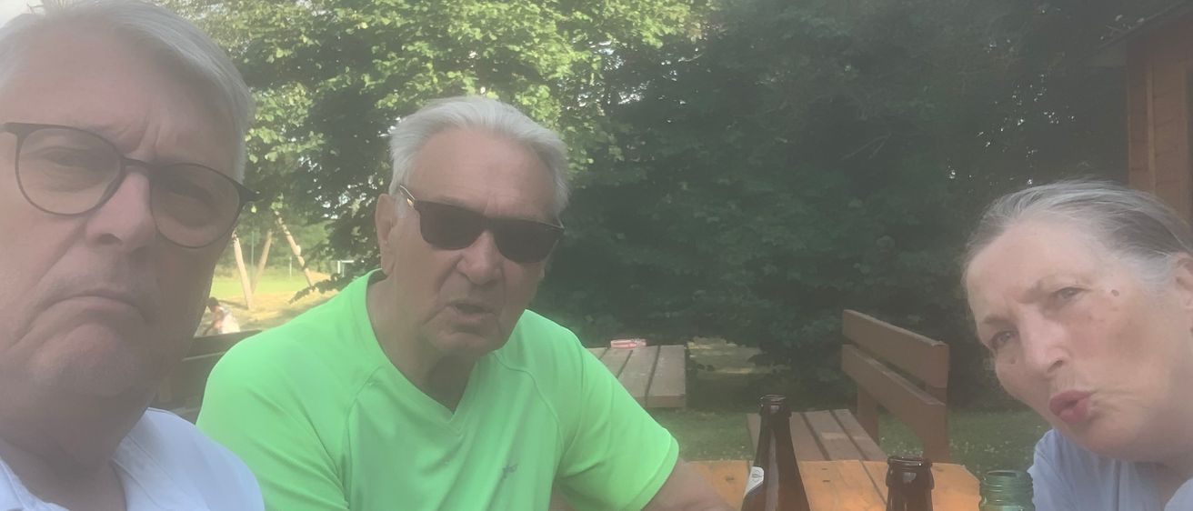 Drei ältere Männer sitzen an einem Picknicktisch mit mehreren Bierflaschen vor ihnen. Der Mann im grünen Shirt hält eine Flasche. Hinter ihnen sind mehr Bänke und Bäume.