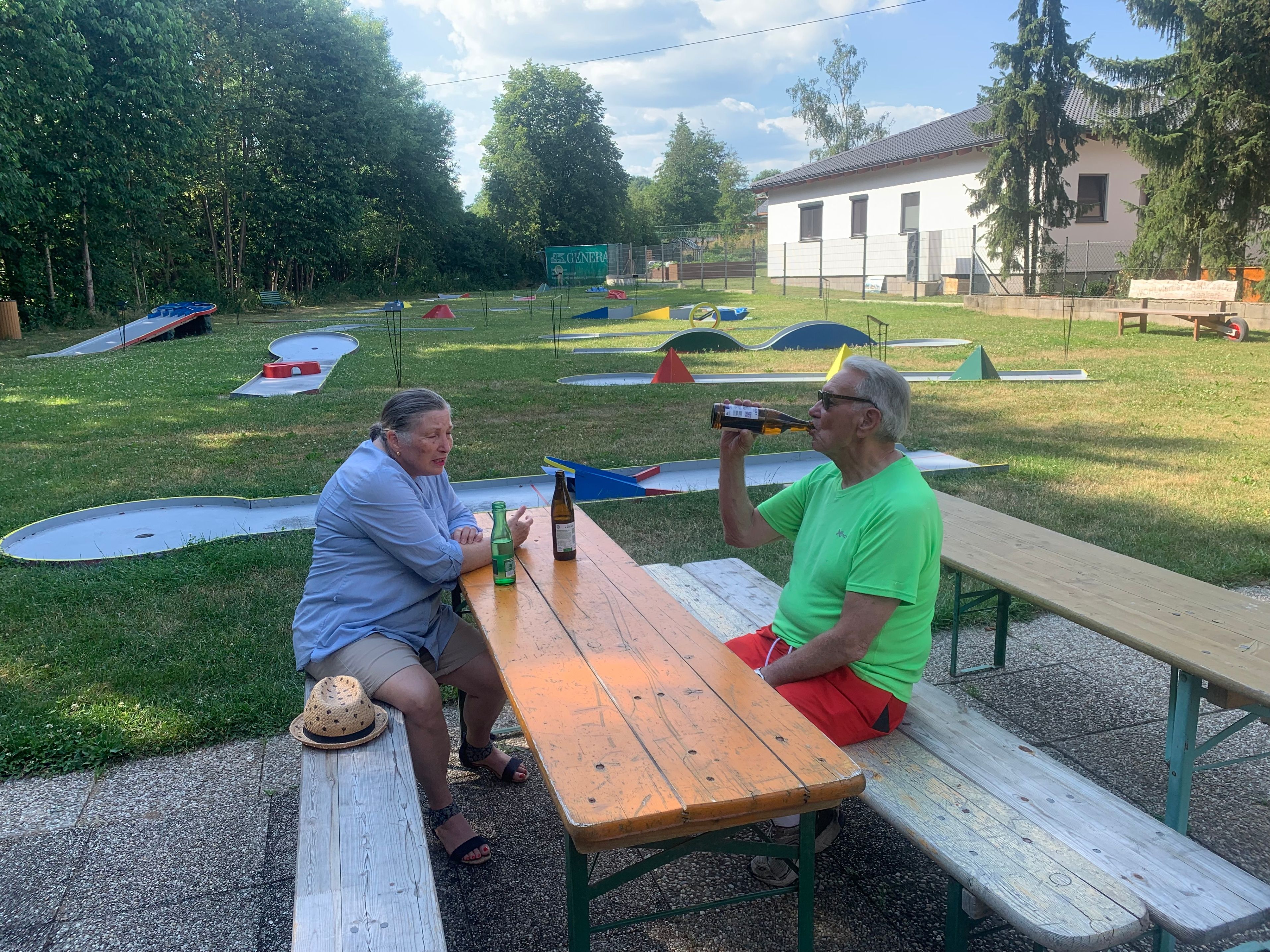 Zwei Männer genießen Getränke auf einer Bank in einem Biergarten. Einer trinkt aus einer Flasche, während der andere zuschaut, mit Minigolfbahnen im Hintergrund.
