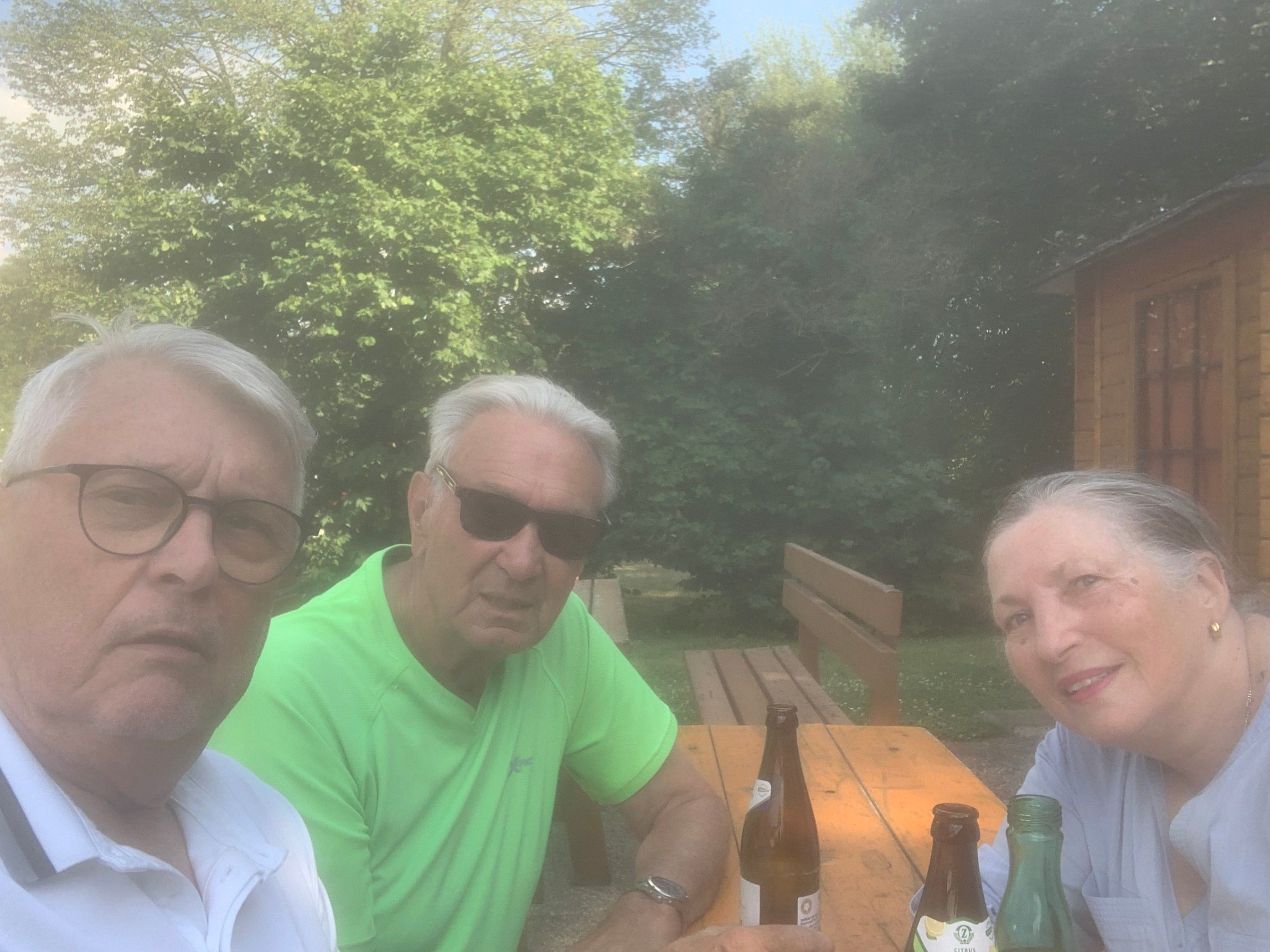 Drei Menschen sitzen lächelnd um einen Holztisch. Dahinter stehen Holzbänke und Bäume. Der Mann links trägt eine Brille und ein weißes Hemd. Der Mann in der Mitte trägt eine Sonnenbrille und ein grünes Hemd. Die Frau rechts trägt ein blaues Hemd.