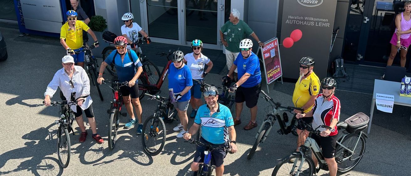 Eine Gruppe von Radfahrern, die Helme und Fahrradausrüstung tragen, steht vor einem Gebäude mit einem Schild, auf dem 'Land Rover' steht. Die Radfahrer posieren für ein Foto.