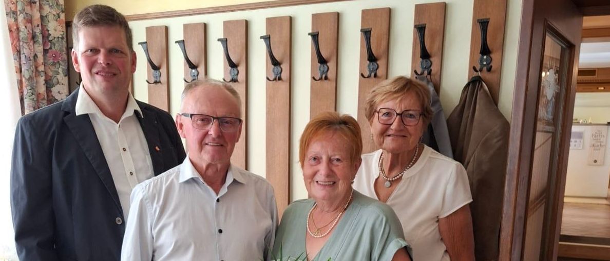 Vier Personen stehen vor einer Tür. Eine Frau hält einen Blumenstrauß. Hinter ihnen sind hölzerne Kleiderhaken. Links steht ein Tisch mit weißer Tischdecke.