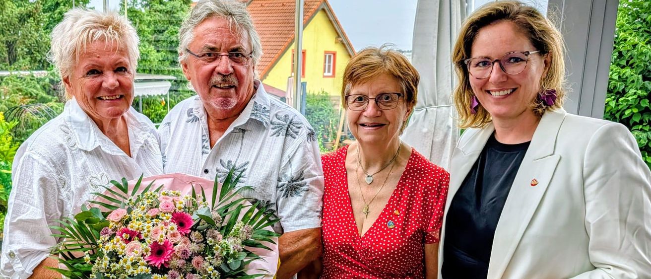 Vier lächelnde Erwachsene stehen zusammen, zwei Männer und zwei Frauen, eine hält einen Geburtstagsstrauß. Sie scheinen einen Geburtstag zu feiern. Hinter ihnen ist ein Haus mit gelber Wand zu sehen.