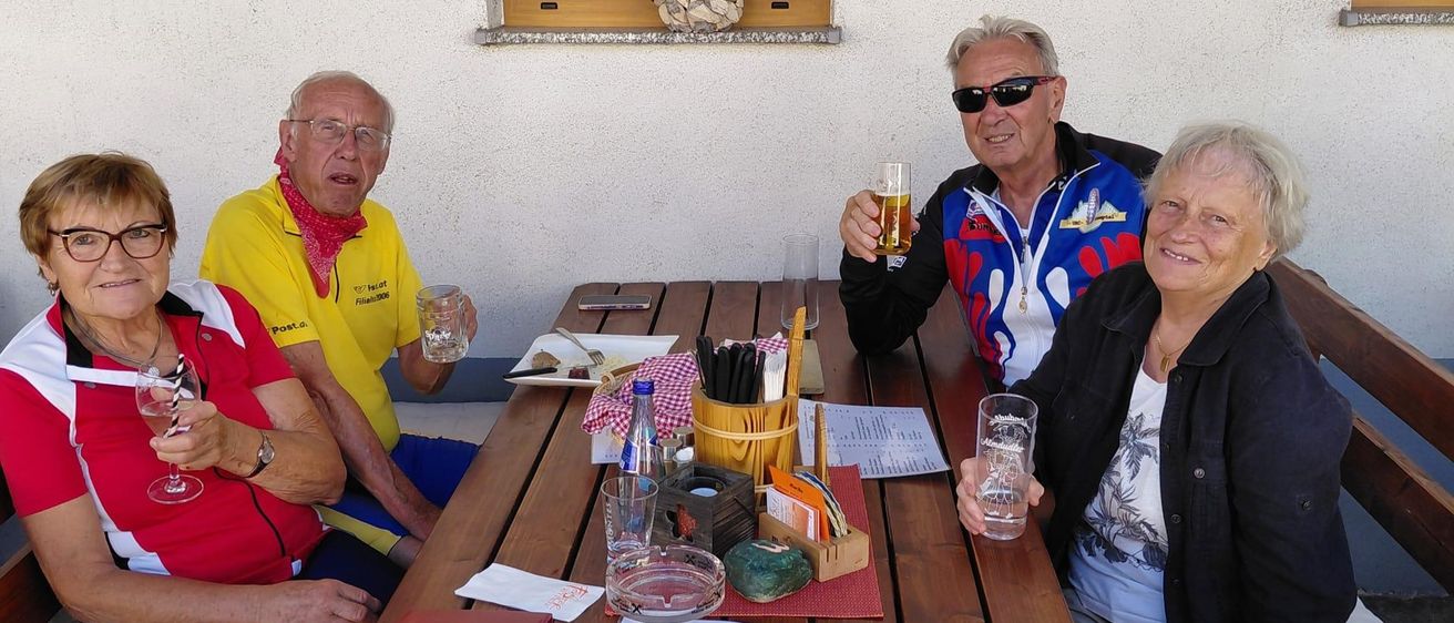 Vier Menschen sitzen an einem Tisch, zwei Männer und zwei Frauen, ein Mann mit Sonnenbrille und gelbem Shirt, der andere in einem blauen Jacket, eine Frau in einem schwarzen Jacket, alle lächeln.