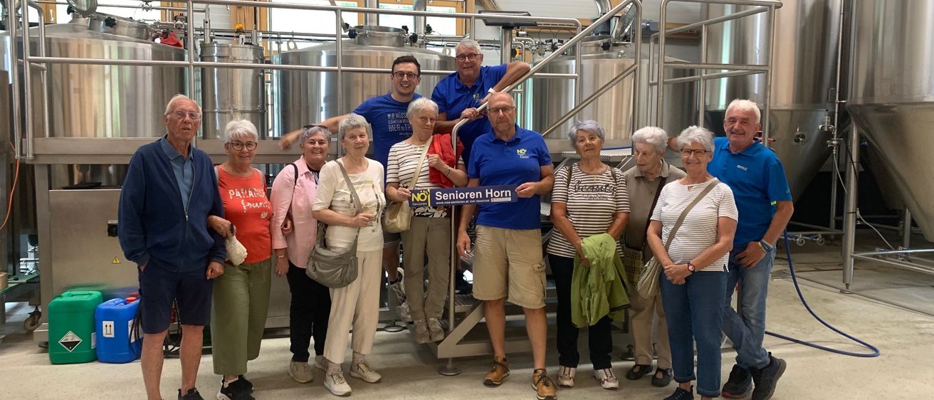 Eine Gruppe von Menschen posiert in einer Brauerei mit Edelstahltanks. Sie tragen Freizeitkleidung. Ein Schild heißt 'Senioren Horn'.