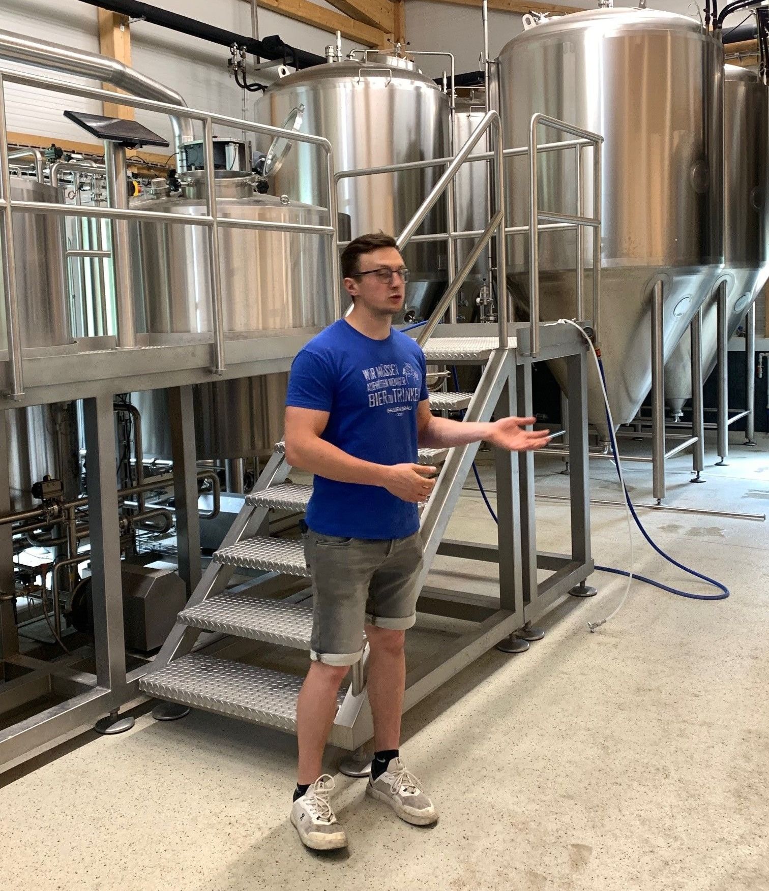 Ein Mann in einem blauen T-Shirt steht in einer Fabrik, spricht, während eine Metalltreppe und große Tanks hinter ihm sind.