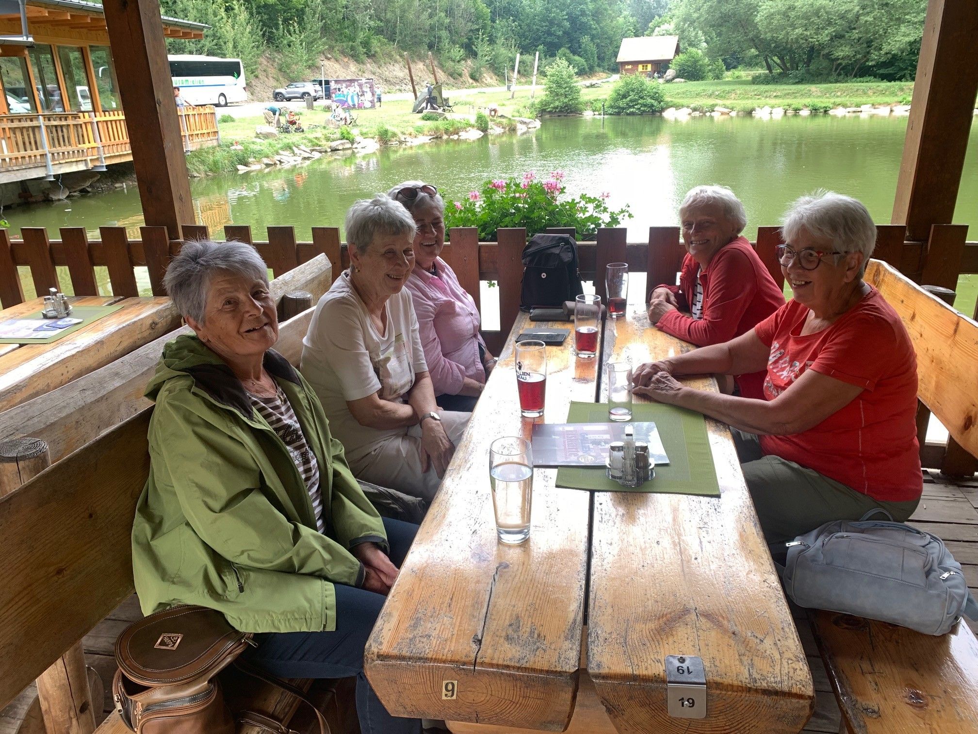 Fünf ältere Frauen sitzen an einem Tisch in einem Restaurant mit einem See im Hintergrund. Sie lächeln und scheinen ihre Mahlzeit zu genießen.