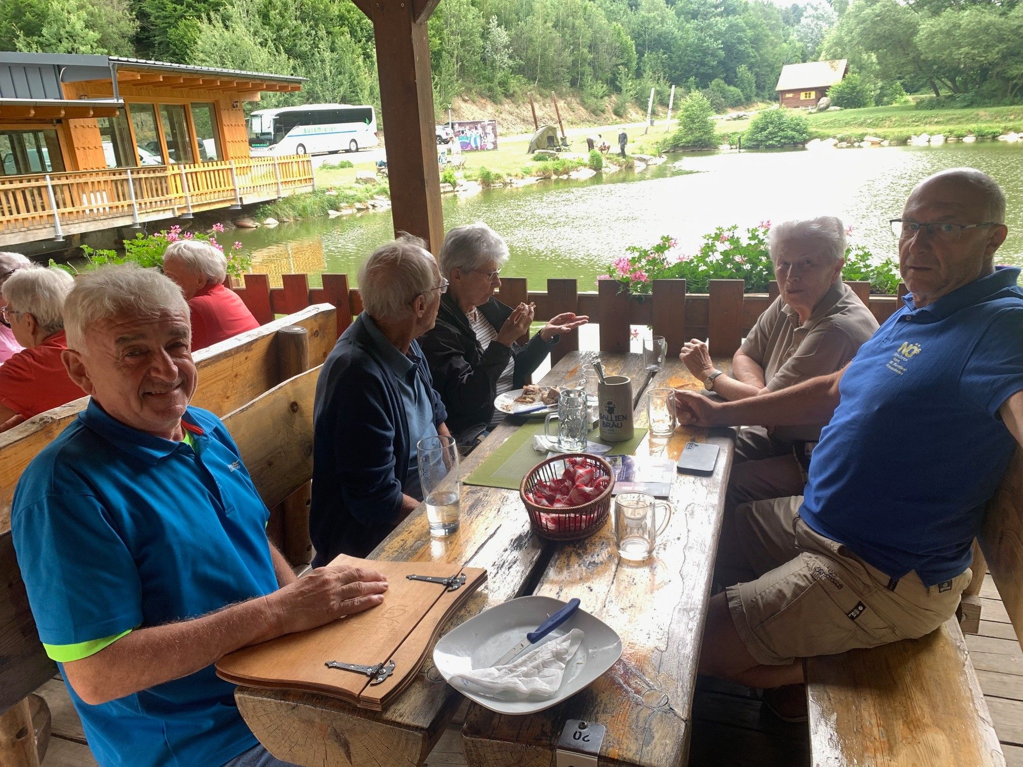 Eine Gruppe älterer Menschen sitzt um einen Holztisch im Freien, in der Nähe eines ruhigen Sees und genießt ihr Essen. Der Tisch ist mit Gläsern, Tellern und einer Körbe mit Süßigkeiten besetzt. Sie sind von einem Holzzaun und einem Blick auf den See umgeben.