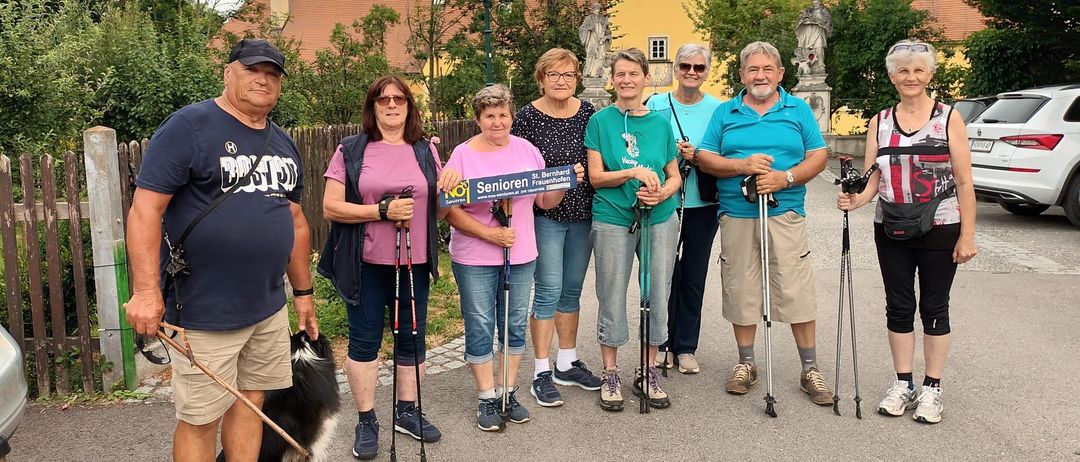 Eine Gruppe von Personen steht im Freien mit Trekkingstöcken, einer hält ein Schild mit Senioren Freiburg. Ein Hund ist bei einem Mann links. Sie stehen vor einem historischen Gebäude.