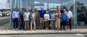 Eine Gruppe von Menschen steht vor einem Sauberer-Autohaus und hält ein Banner. Sie lächeln und scheinen für ein Foto zu posieren. Dahinter sind Autos auf dem Gelände geparkt.