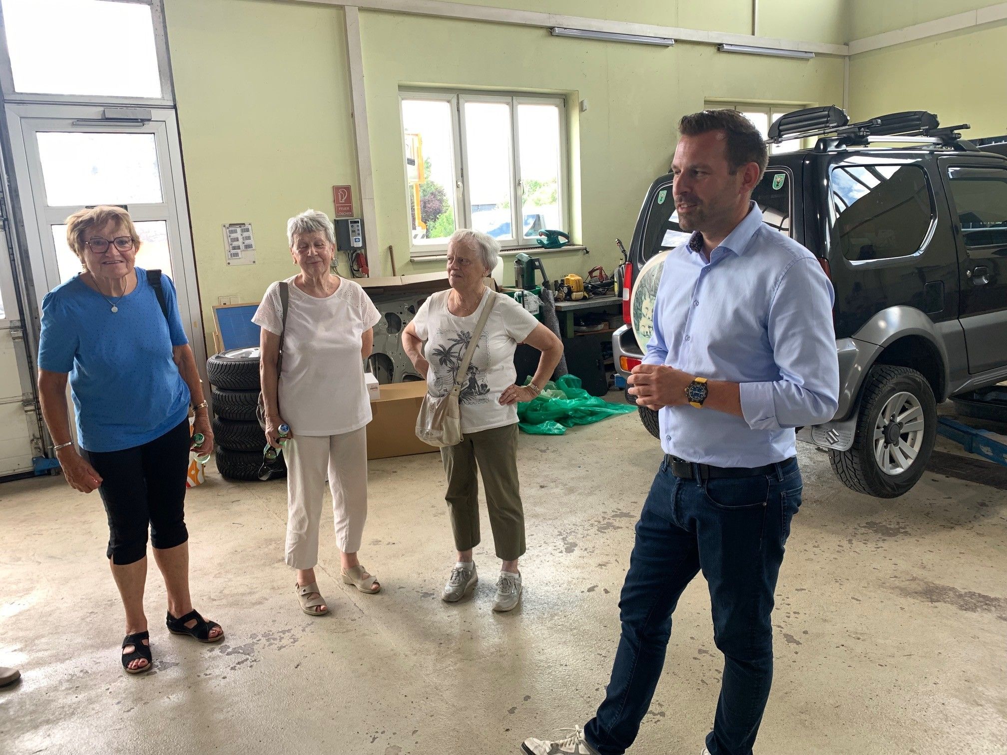 Vier Personen stehen in einer Garage, zwei ältere Frauen in weißen Shirts und ein Mann in einem hellblauen Hemd. Der Mann scheint etwas zu erklären.