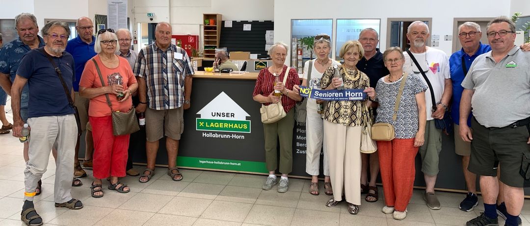 Eine Gruppe von Menschen, hauptsächlich Senioren, steht in einem Geschäft zusammen. Sie lächeln und einige halten Getränke. Ein Schild hinter ihnen sagt 'Senioren Horn'.