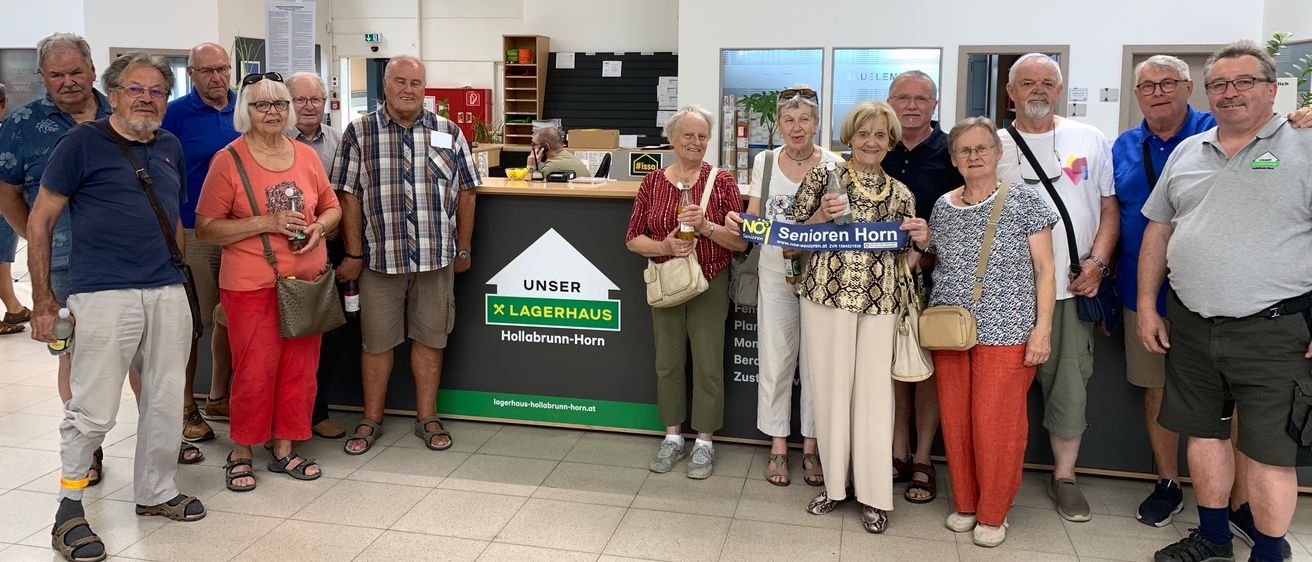 Eine Gruppe von Menschen, hauptsächlich Senioren, steht in einem Geschäft zusammen. Sie lächeln und einige halten Getränke. Ein Schild hinter ihnen sagt 'Senioren Horn'.