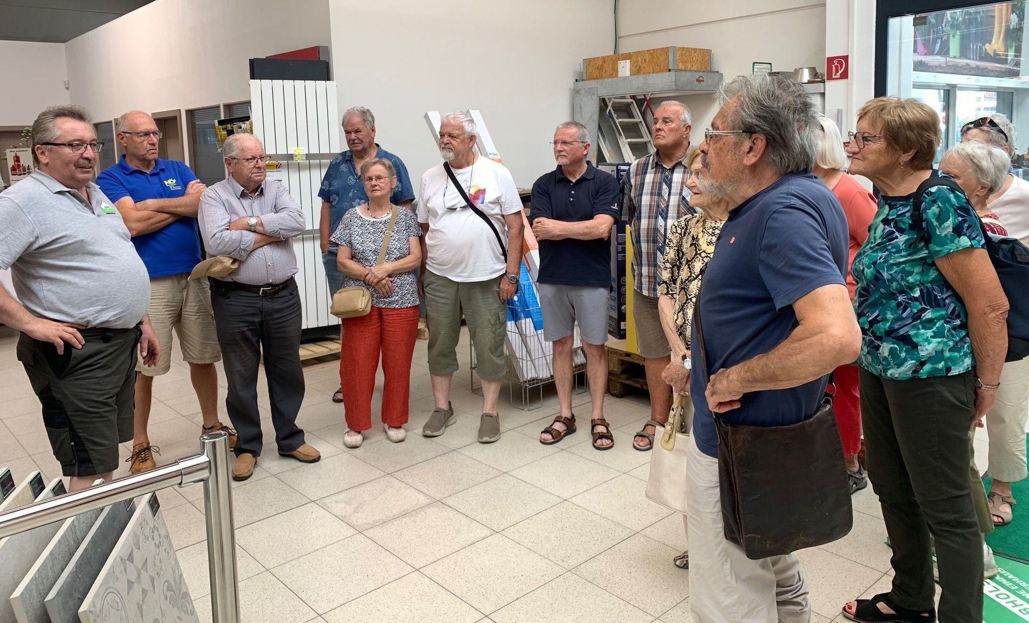 Eine Gruppe älterer Erwachsener steht in einem gefliesten Raum, einige tragen Brillen, halten Taschen und einer eine Uhr. Sie scheinen zu sprechen.