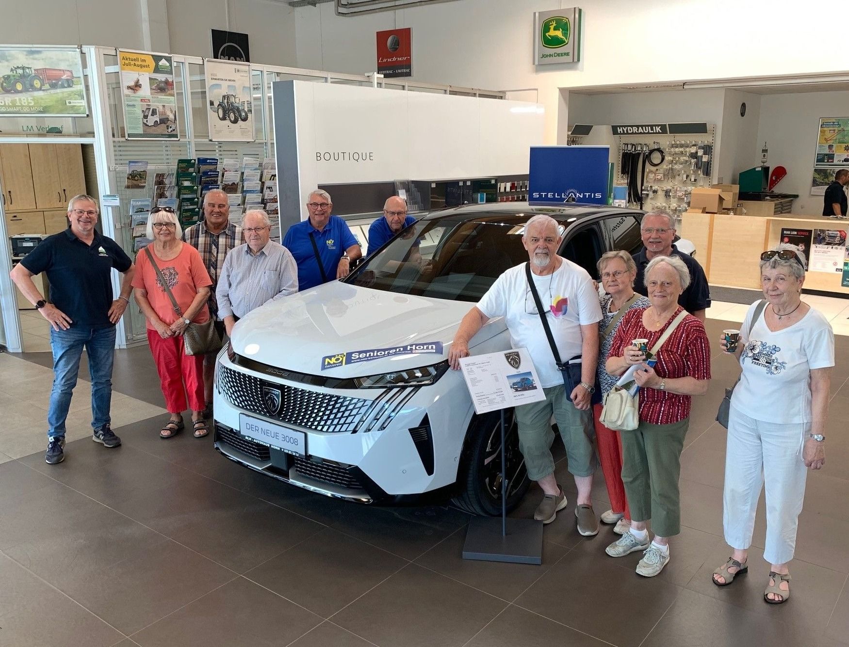 Eine Gruppe von Menschen steht neben einem weißen Auto in einer Ausstellung. Sie scheinen es zu bewundern. Sie alle lächeln.