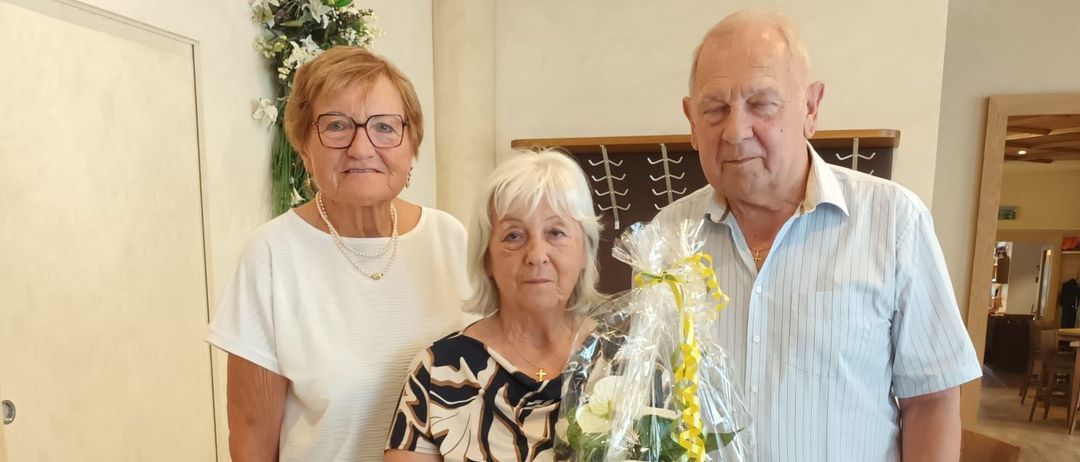 Drei ältere Personen stehen zusammen in einem Raum, wobei eine Frau einen Blumenstrauß hält. Die Frau auf der linken Seite trägt eine Brille und eine Halskette. Der Mann auf der rechten Seite trägt einen Gürtel und eine Uhr.