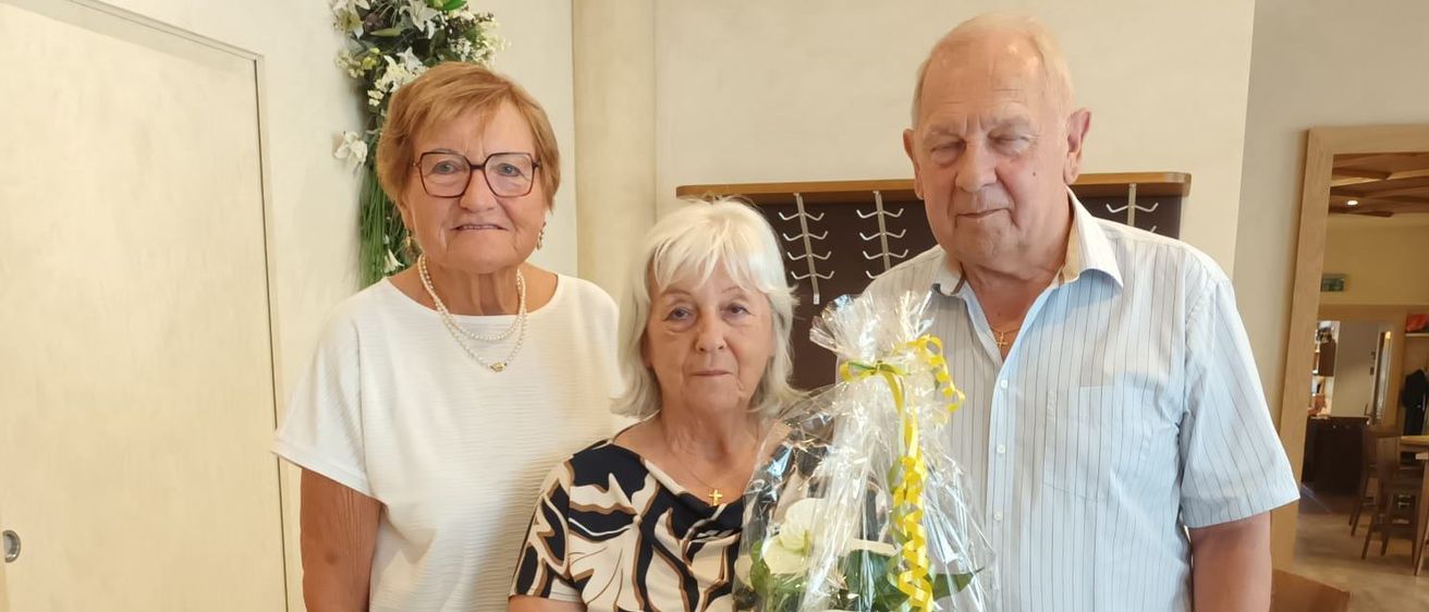 Drei ältere Personen stehen zusammen in einem Raum, wobei eine Frau einen Blumenstrauß hält. Die Frau auf der linken Seite trägt eine Brille und eine Halskette. Der Mann auf der rechten Seite trägt einen Gürtel und eine Uhr.