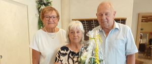 Drei ältere Personen stehen zusammen in einem Raum, wobei eine Frau einen Blumenstrauß hält. Die Frau auf der linken Seite trägt eine Brille und eine Halskette. Der Mann auf der rechten Seite trägt einen Gürtel und eine Uhr.