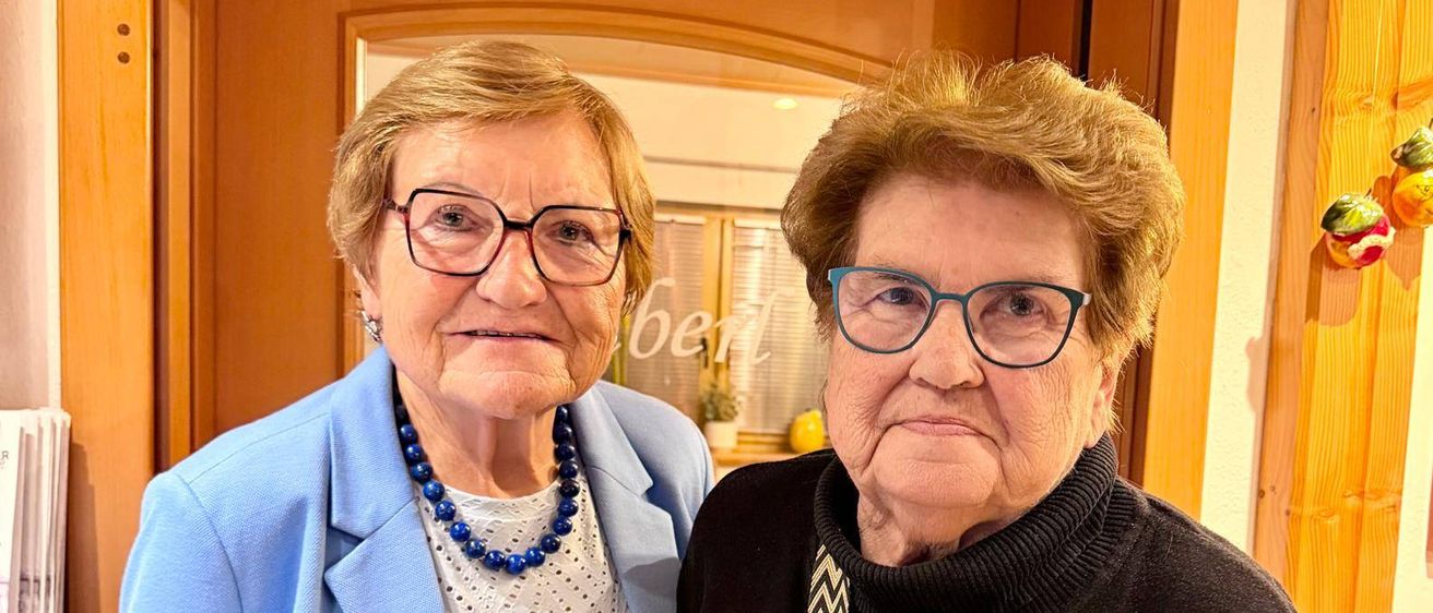 Zwei ältere Frauen stehen vor einer Holztür. Die Frau auf der linken Seite trägt einen blauen Blazer und eine Halskette. Die Frau auf der rechten Seite hält einen Strauß roter Blumen und trägt ein schwarzes Rollkragenshirt mit Leopardenmuster.