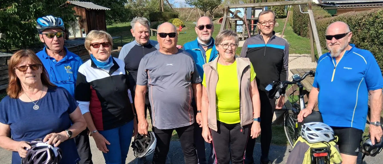 Eine Gruppe von Menschen steht zusammen in einem Park, alle tragen Fahrradausrüstung und halten Helme. Sie lächeln und scheinen für ein Foto zu posieren.