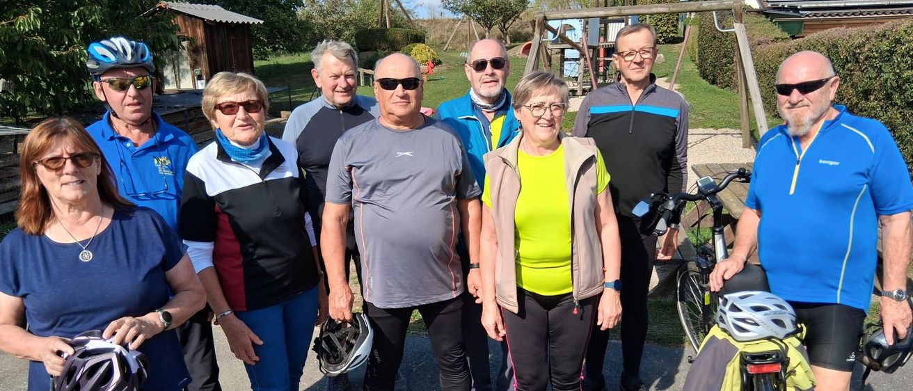 Eine Gruppe von Menschen steht zusammen in einem Park, alle tragen Fahrradausrüstung und halten Helme. Sie lächeln und scheinen für ein Foto zu posieren.