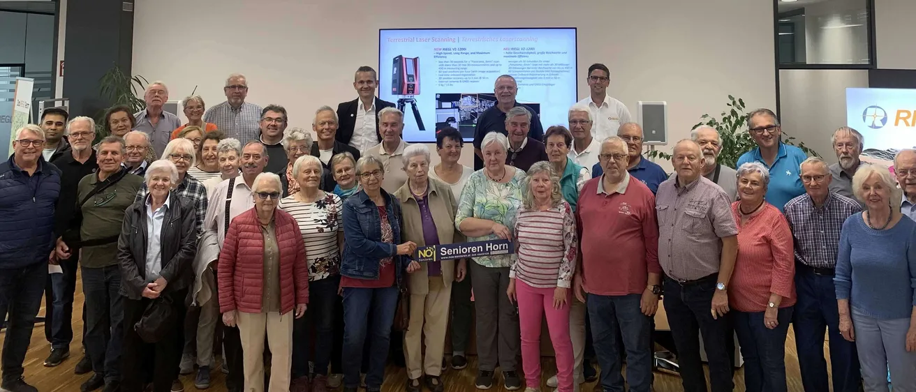 Eine Gruppe von Menschen, hauptsächlich Senioren, ist in einem Raum für ein Foto versammelt. Einige tragen Brillen und einige tragen Jacken. Eine Projektionsleinwand befindet sich an der Wand hinter ihnen, und es gibt Banner mit dem Text 'RIEGL' auf beiden Seiten.