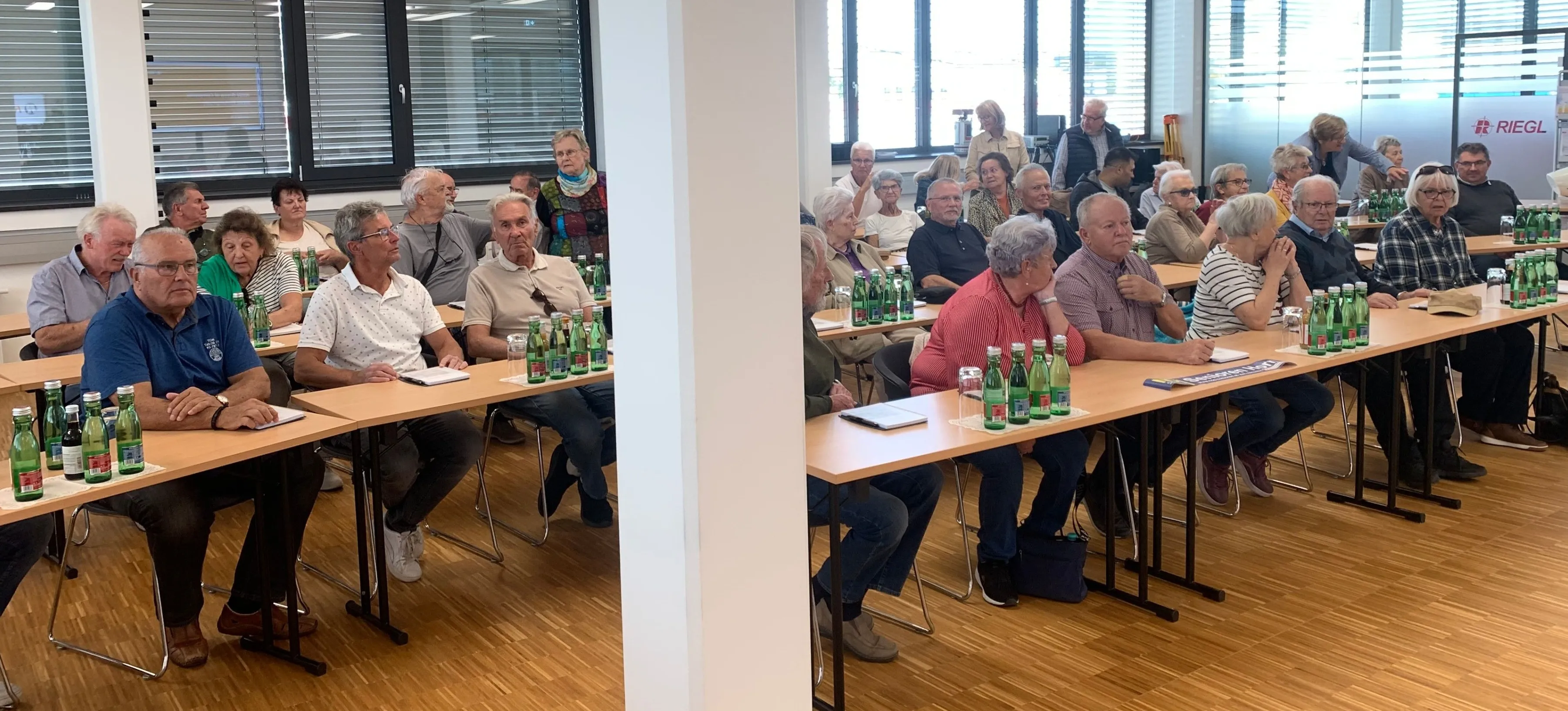 Eine Gruppe von Menschen sitzt in einem Konferenzraum und nimmt an einer Besprechung teil. Der Raum hat Holzböden und große Fenster.