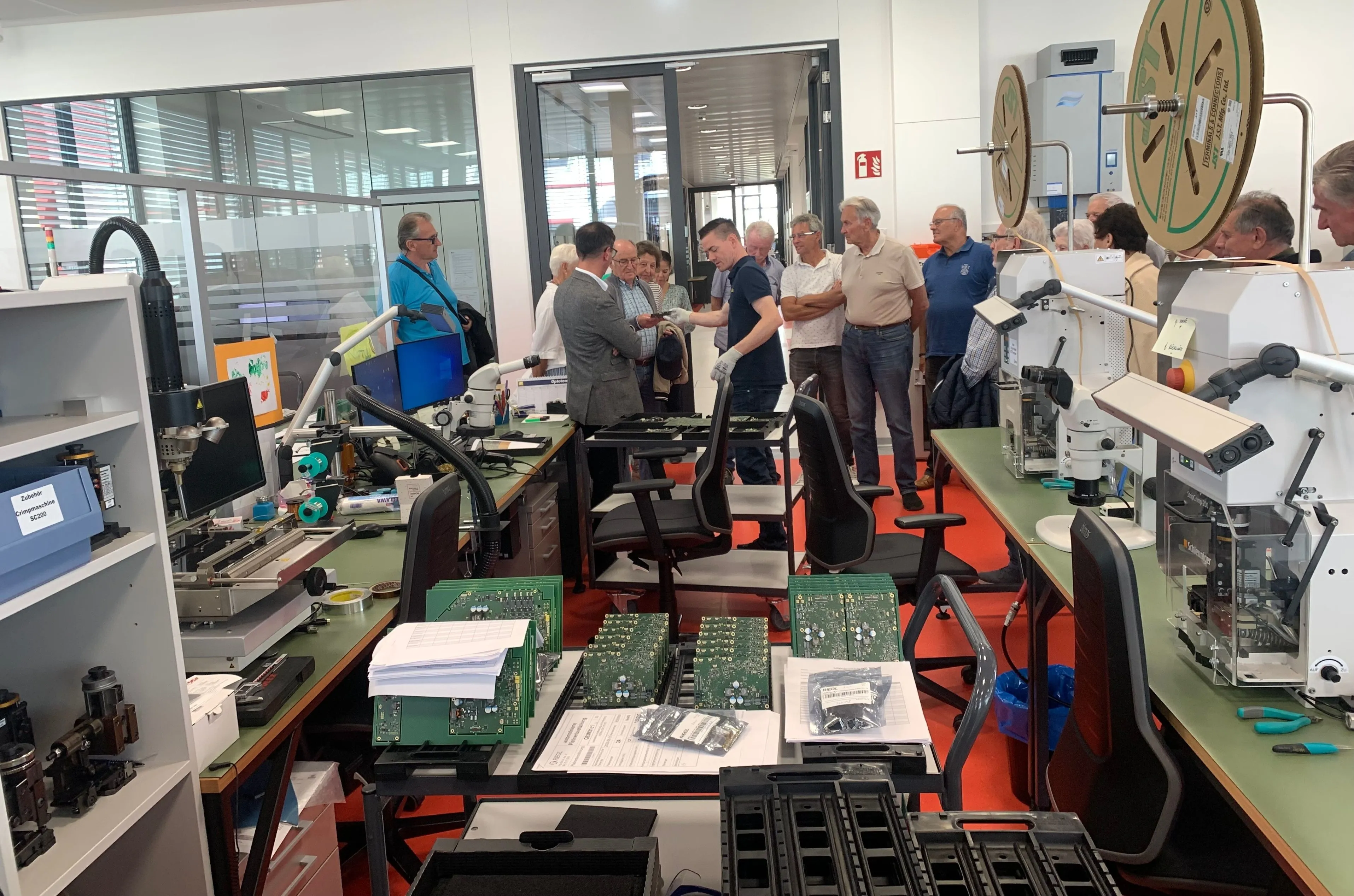 Eine Gruppe von Menschen steht in einer Werkstatt und betrachtet elektronische Komponenten auf einem Tisch.