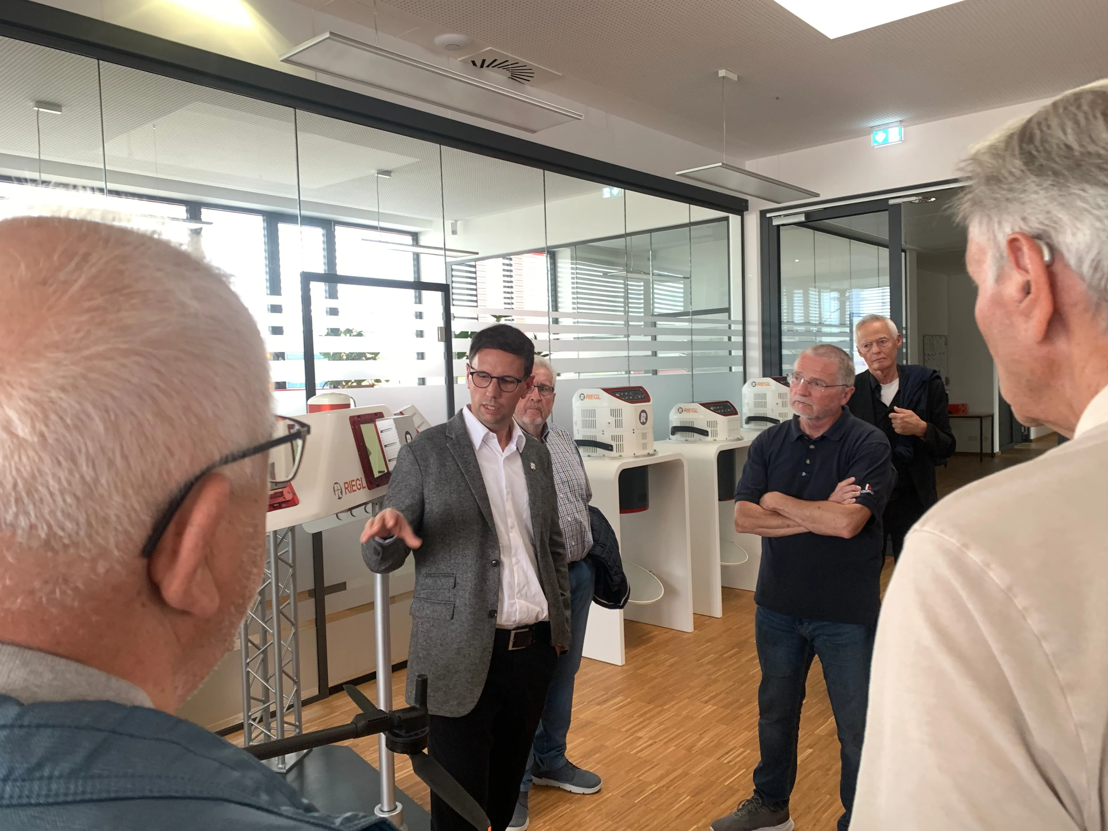 Eine Gruppe von Männern versammelt sich in einem Raum mit Glaswänden. Ein Mann spricht mit einem anderen Mann, während die anderen zuhören.