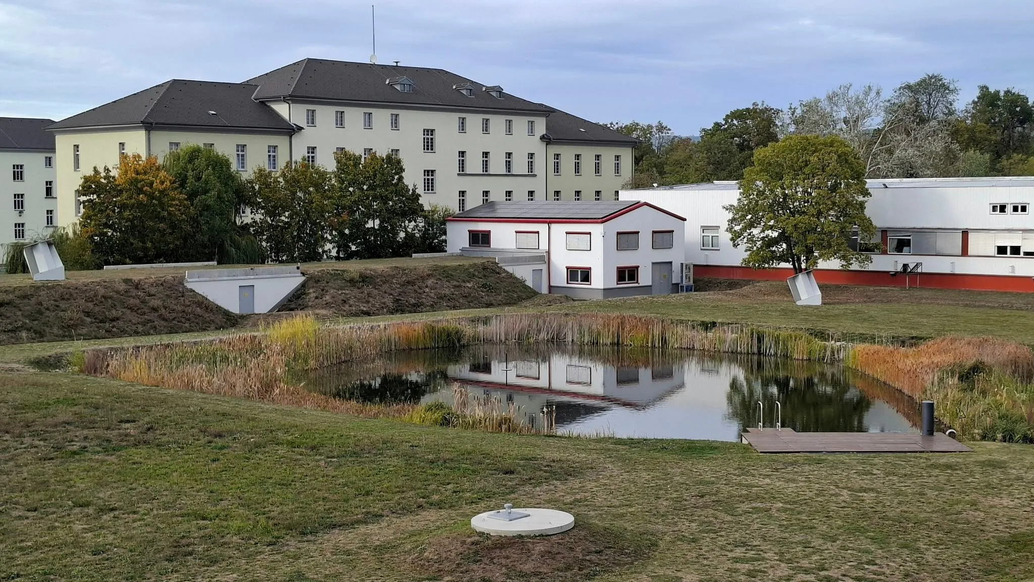 Ein großes Gebäude mit grauem Dach spiegelt sich in einem Teich vor ihm.
