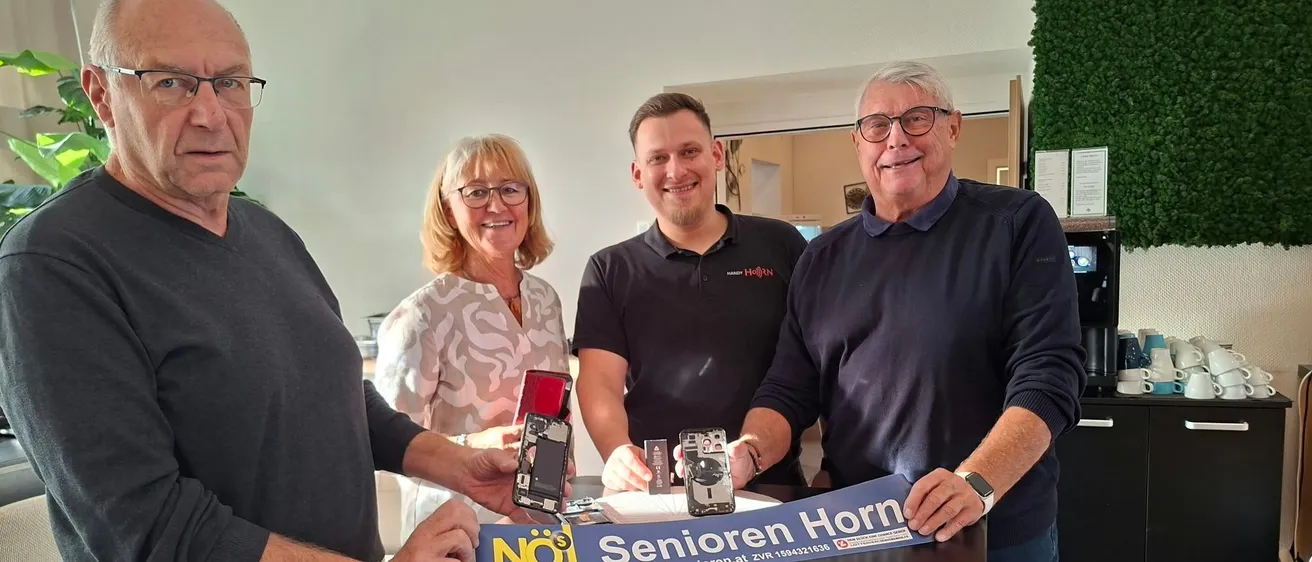 Vier Personen stehen um einen Tisch herum, zwei Männer und zwei Frauen. Sie halten ein Banner und einige Gegenstände.