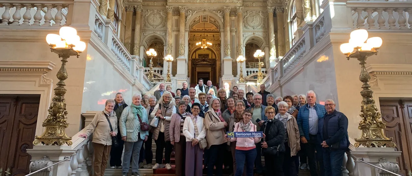 Eine Gruppe von Menschen steht auf den Treppen eines großen Gebäudes und posiert für ein Foto. Die Treppen sind mit rotem Teppich ausgelegt und haben kunstvolle Lampen.