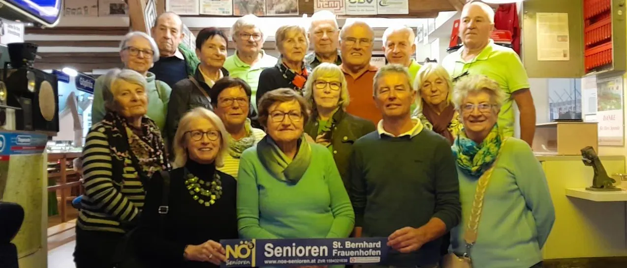 Eine Gruppe älterer Menschen steht in einem Raum, lächelt und posiert für ein Foto. Sie tragen grüne Kleidung und halten ein Schild mit der Aufschrift 'Senioren' und 'St. Bernhard Frauenhofen'.