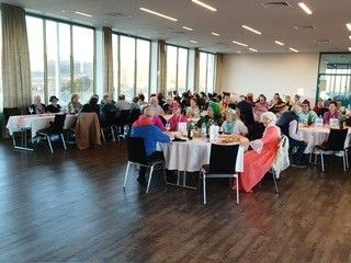 Ein großer Raum mit Holzböden und großen Fenstern zeigt eine Gruppe von Menschen, die an Tischen sitzen, einige mit Kopfbedeckungen, mit Essen und Wein auf den Tischen.