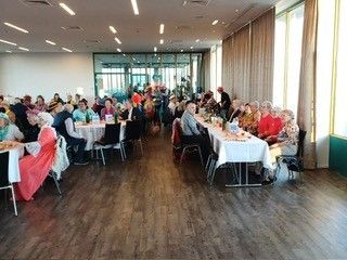 Ein großer Raum mit vielen Menschen, die an Tischen mit weißen Tischdecken sitzen. Der Raum hat Holzböden, Glasfenster und hohe Decken.