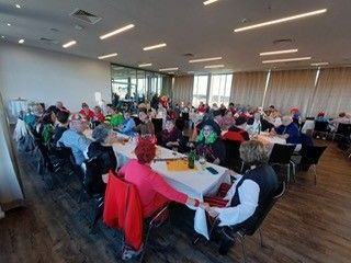 Eine Gruppe von Menschen in verschiedenen Kostümen sitzt an langen Tischen in einem gut beleuchteten Bankettsaal mit großen Fenstern und Holzböden.