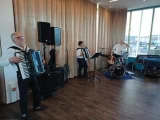 Drei Männer spielen Instrumente in einem Raum mit Holzböden und einem großen Fenster. Einer spielt Akkordeon, ein anderer Schlagzeug und der dritte Gitarre.